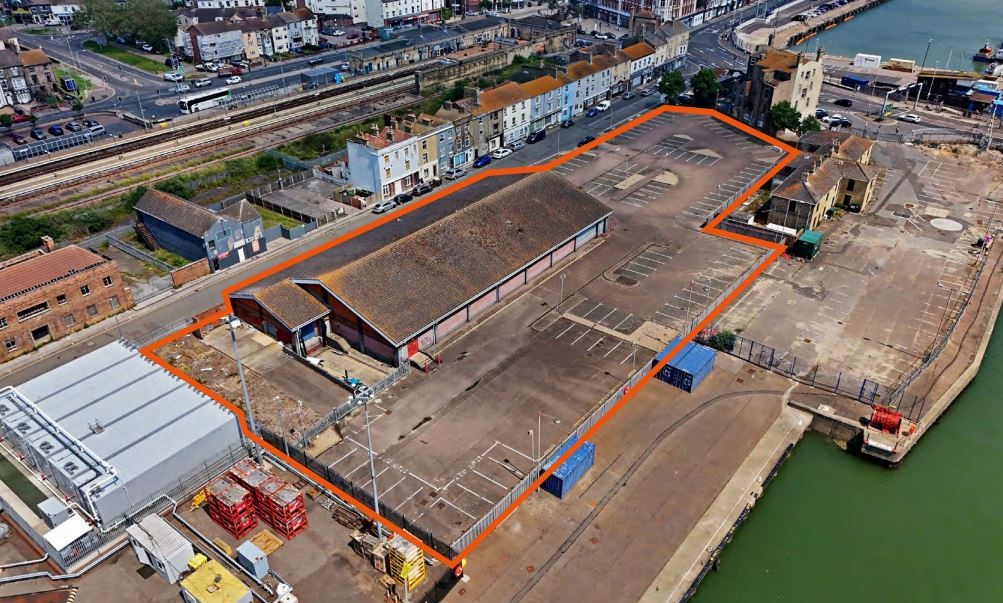 Oblique aerial photo of site including a large building surrounded by a carpark, with perimeter of area outlined in orange