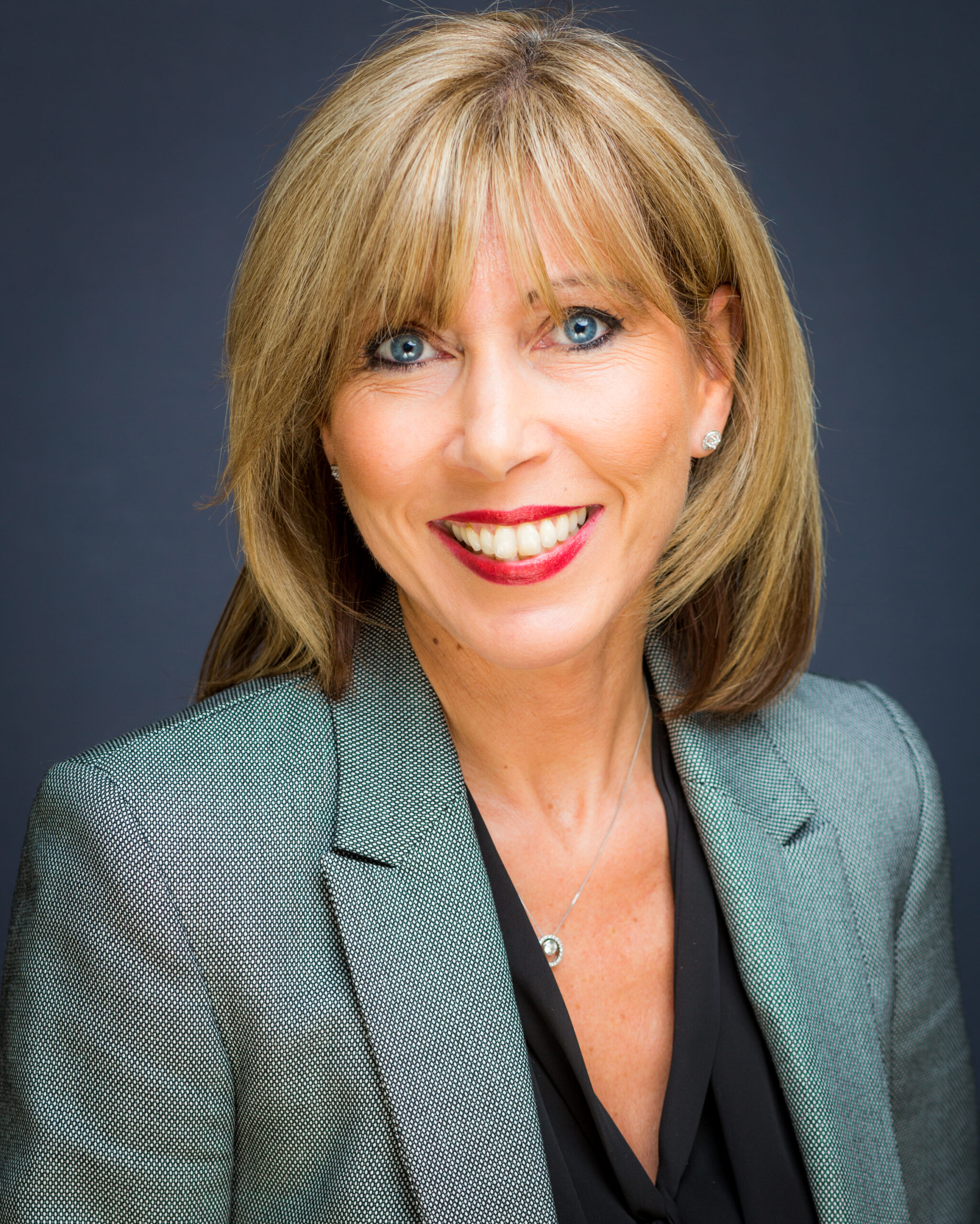 A person with shoulder-length blonde hair is wearing a grey blazer over a black top. The background is a plain dark color
